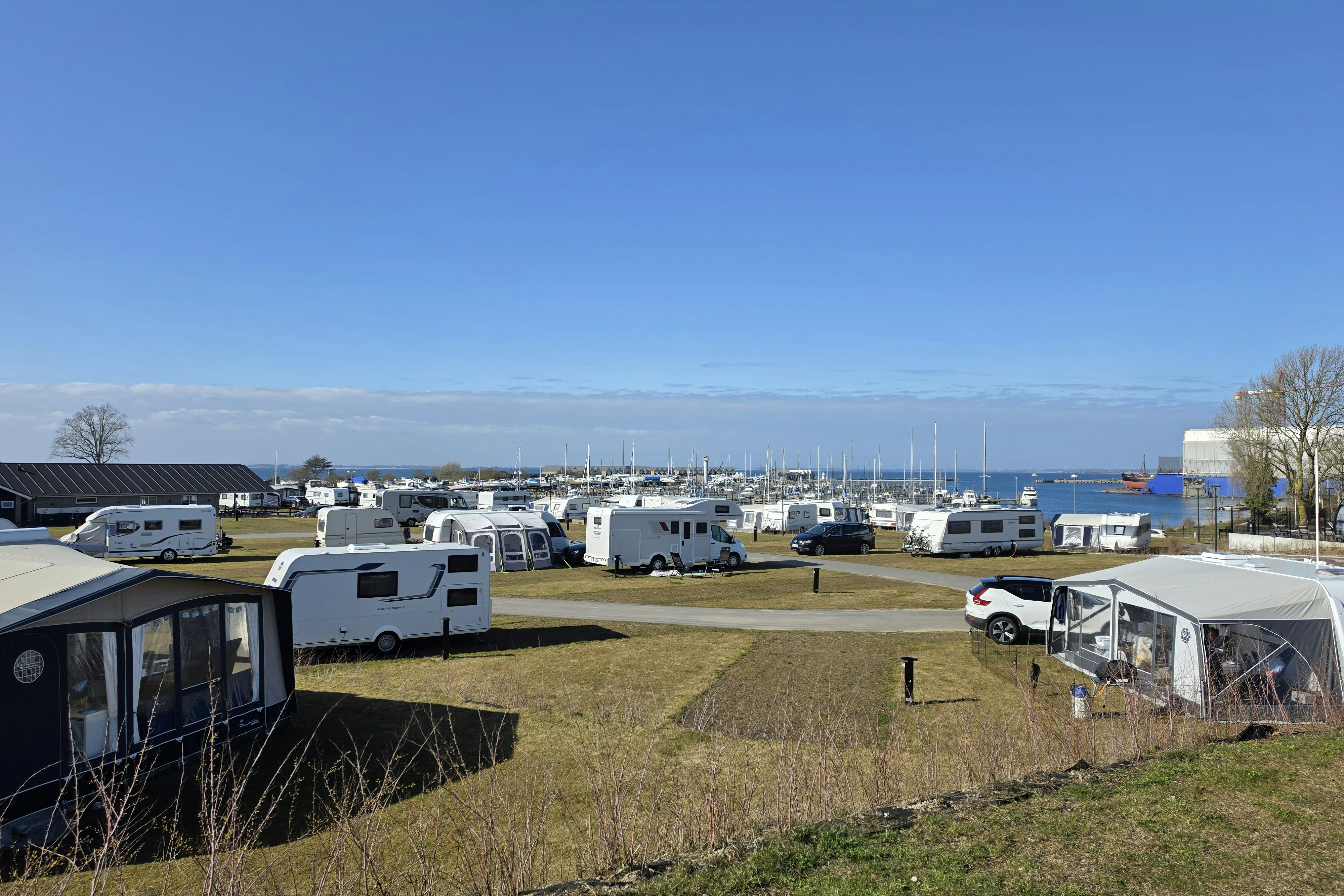 Wohnwagen und Zelte auf einem Campingplatz mit Stellplätzen, im Hintergrund Hafen mit vielen Booten und Meer.