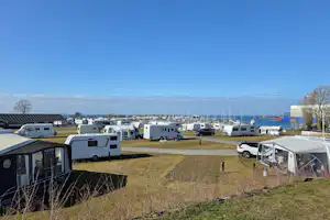 Wohnwagen und Zelte auf einem Campingplatz mit Stellplätzen, im Hintergrund Hafen mit vielen Booten und Meer.