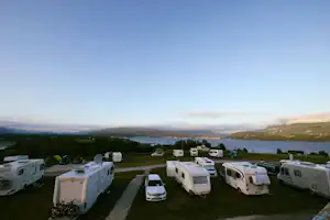 Wohnmobile und Autos auf einem Campingplatz am See, im Hintergrund bewaldete Hügel und eine weite Bucht.