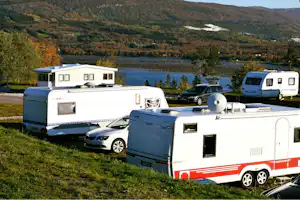 Wohnmobile und Autos auf einem Campingplatz am See, im Hintergrund bewaldete Hügel und ein Tal.