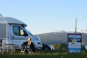 Wohnmobil auf einem Campingplatz mit Leiter, im Hintergrund Berge und ein Schild mit Camping-Informationen.