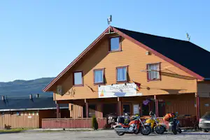 Gelbes Holzhaus mit Balkon und Schildern, davor mehrere Motorräder auf dem Parkplatz, im Hintergrund Hügel und Himmel.