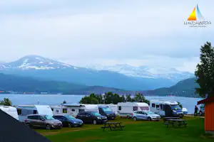 Campingplatz mit Wohnwagen und Autos, Blick auf einen See und bewaldete Berge in der Ferne.