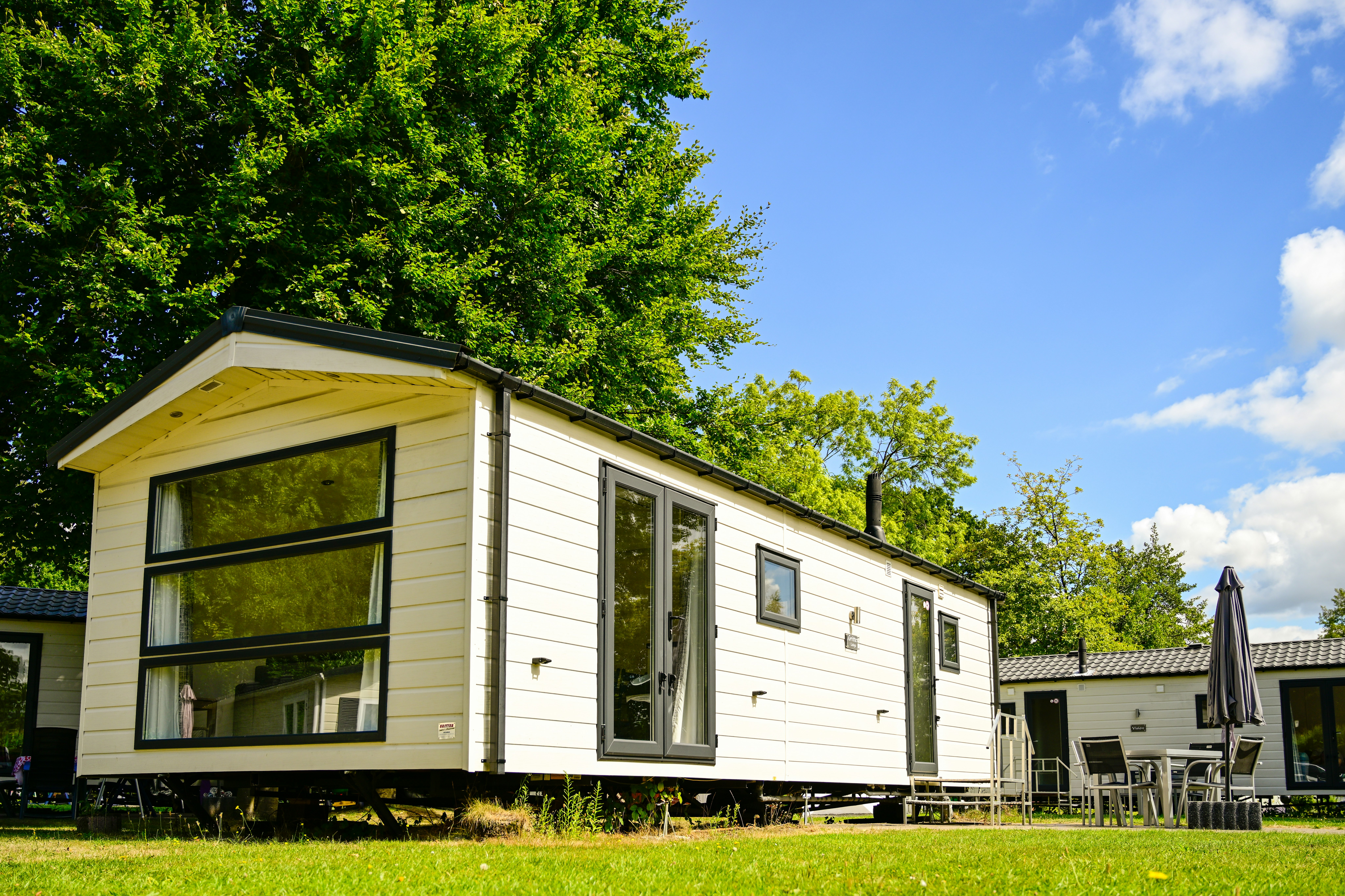 Mobilheim mit großen Fenstern und Terrasse am Campingplatz, umgeben von Rasen und Bäumen.
