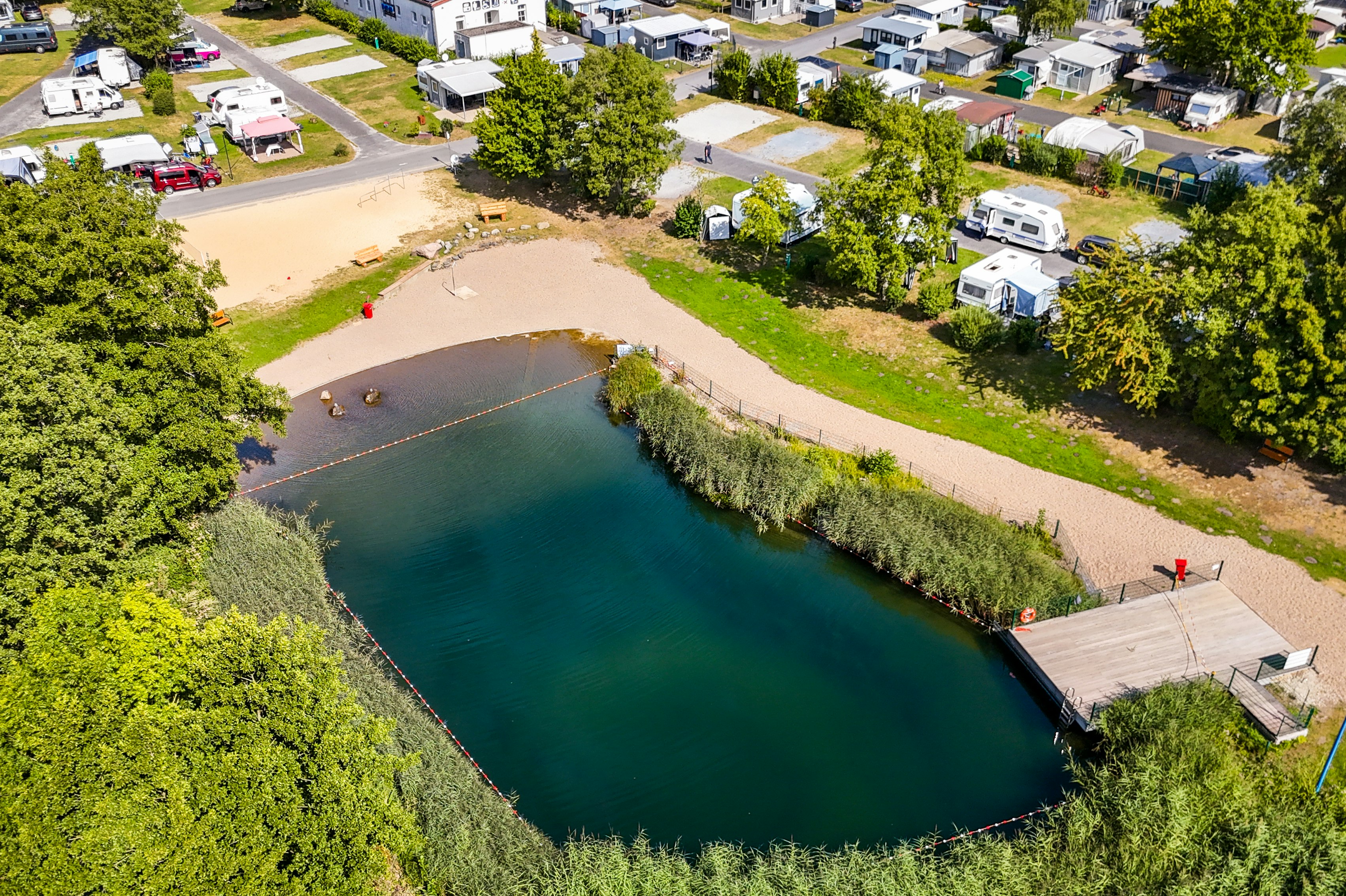 Blick auf den See mit Sandstrand, Steg und Wohnmobilen auf Stellplätzen, umgeben von Bäumen und Wegen.