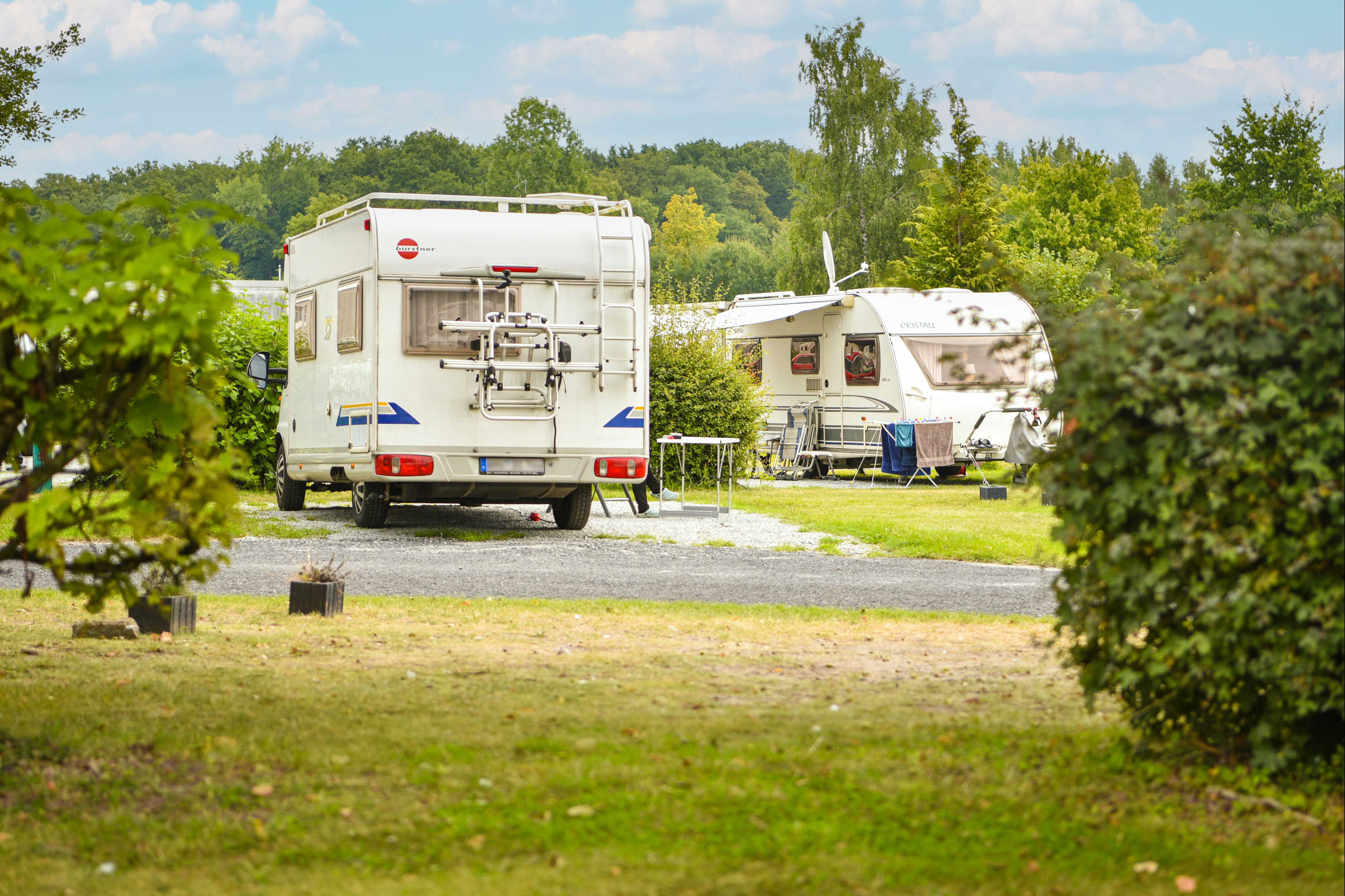 Zwei Wohnmobile auf einem Campingplatz, Stellplatz mit Gras und Kiesweg, umgeben von Bäumen und Sträuchern.