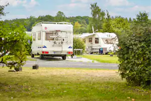 Zwei Wohnmobile auf einem Campingplatz, Stellplatz mit Gras und Kiesweg, umgeben von Bäumen und Sträuchern.