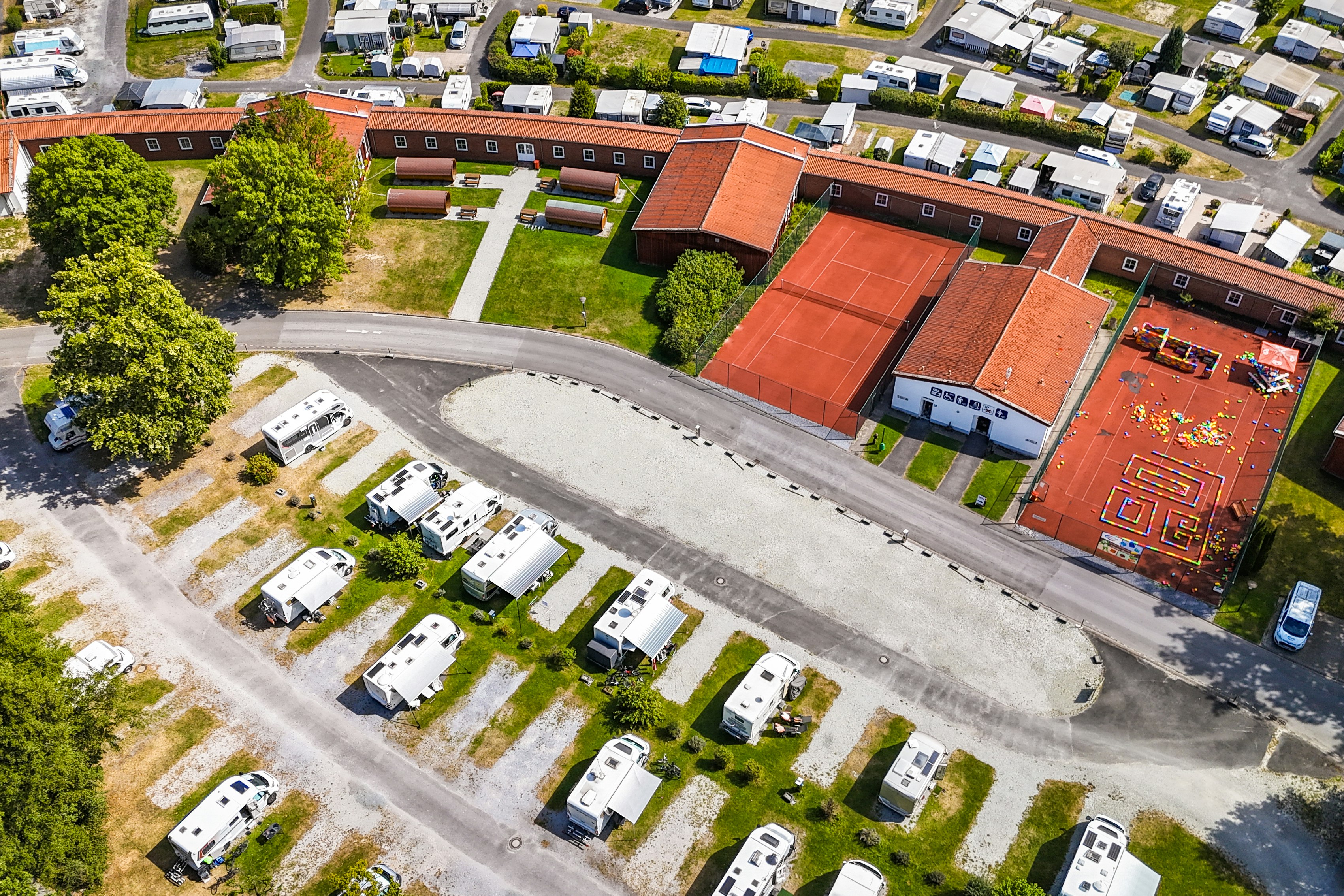 Luftaufnahme eines Campingplatzes mit Wohnwagen, Stellplätzen und Wegen, umgeben von Bäumen und mehreren Gebäuden mit roten Dächern.