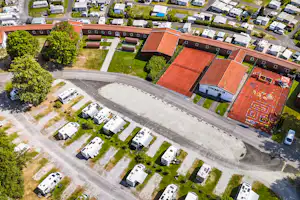 Luftaufnahme eines Campingplatzes mit Wohnwagen, Stellplätzen und Wegen, umgeben von Bäumen und mehreren Gebäuden mit roten Dächern.