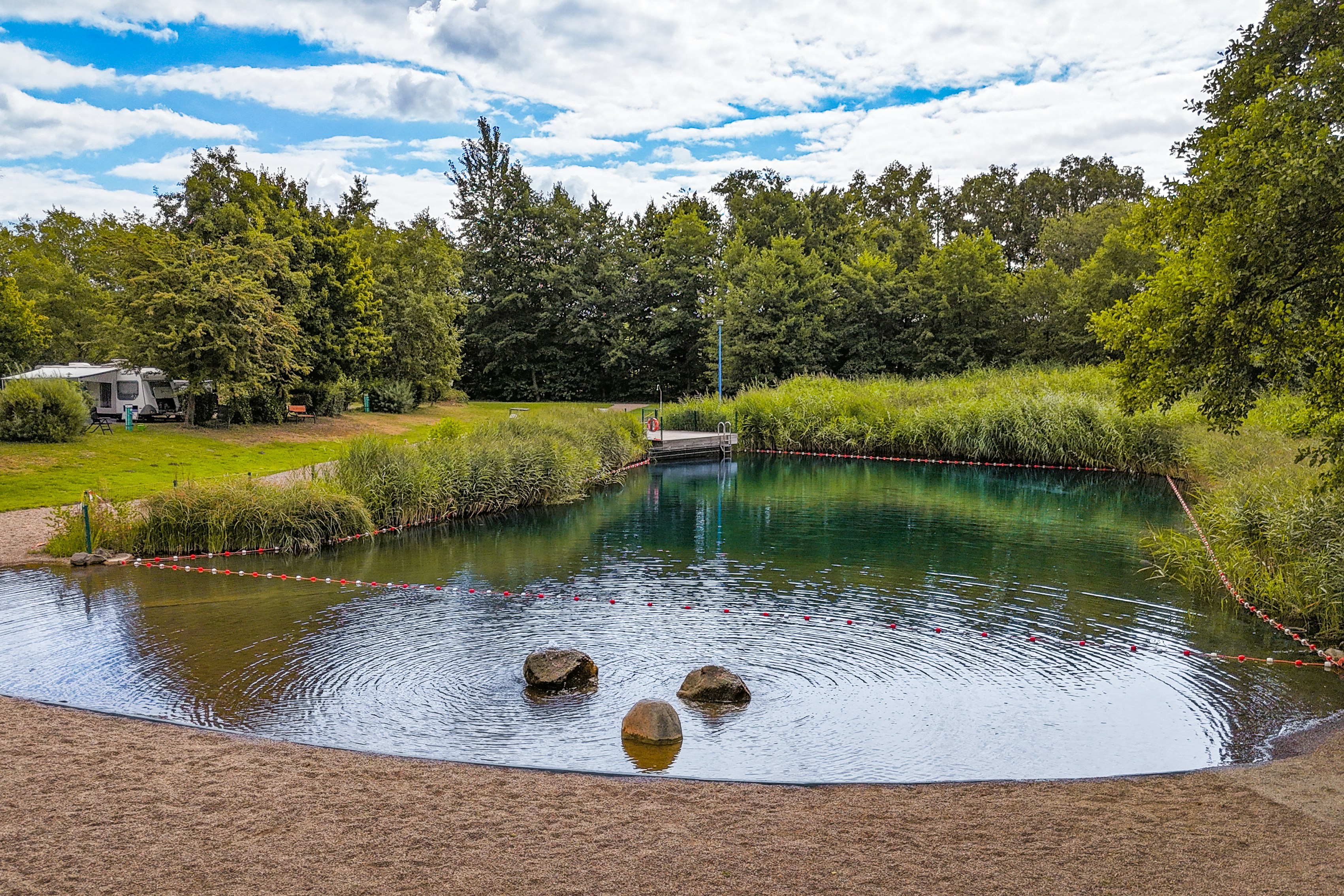 Ruhiger See auf dem Campingplatz mit Steg, Uferweg und Bäumen, im Hintergrund Wohnwagen und Wohnmobilstellplätze.