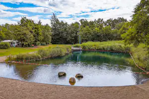Ruhiger See auf dem Campingplatz mit Steg, Uferweg und Bäumen, im Hintergrund Wohnwagen und Wohnmobilstellplätze.