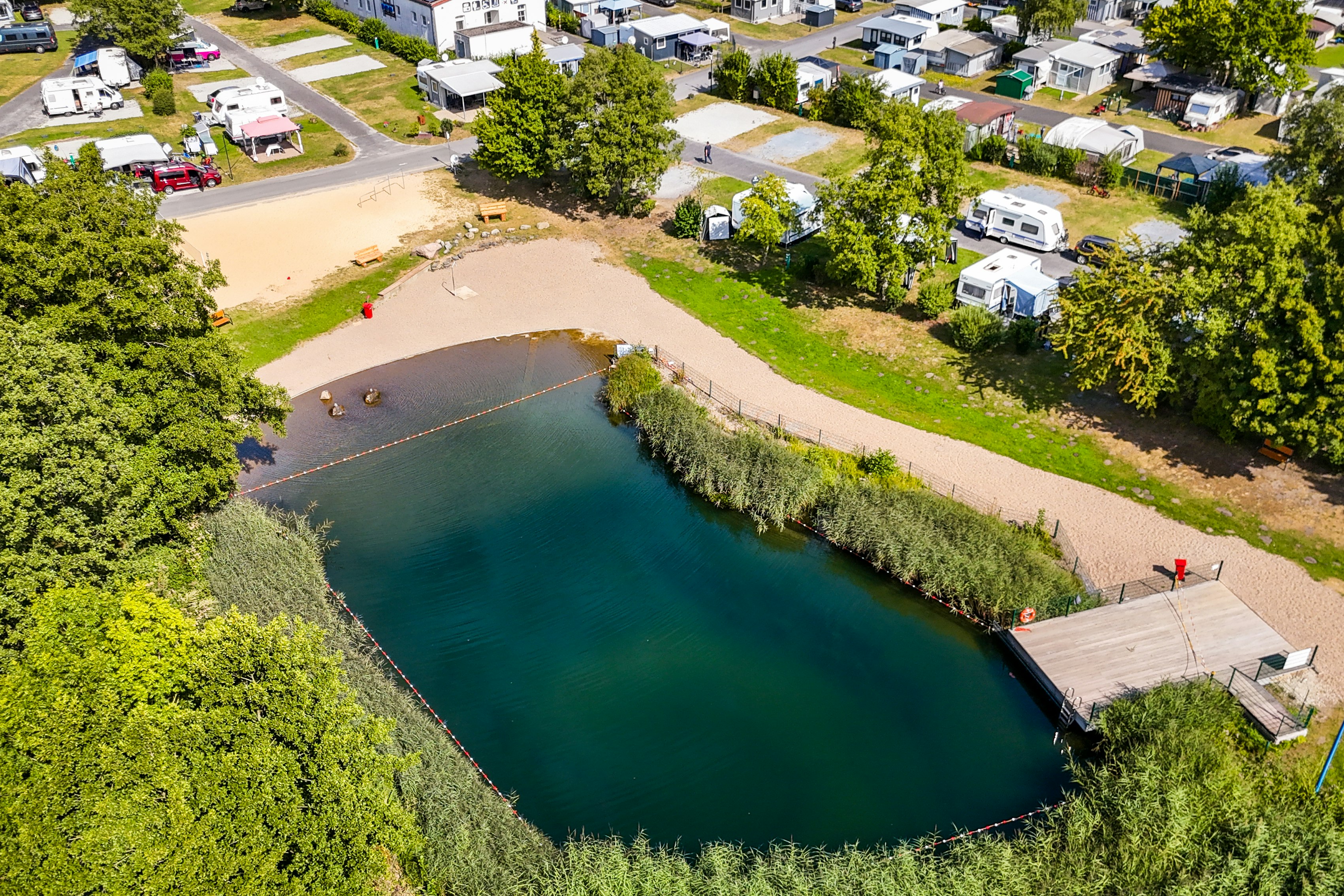 Luftaufnahme eines Campingplatzes am See mit Holzsteg, Uferweg und Stellplätzen mit Wohnwagen und Bäumen.