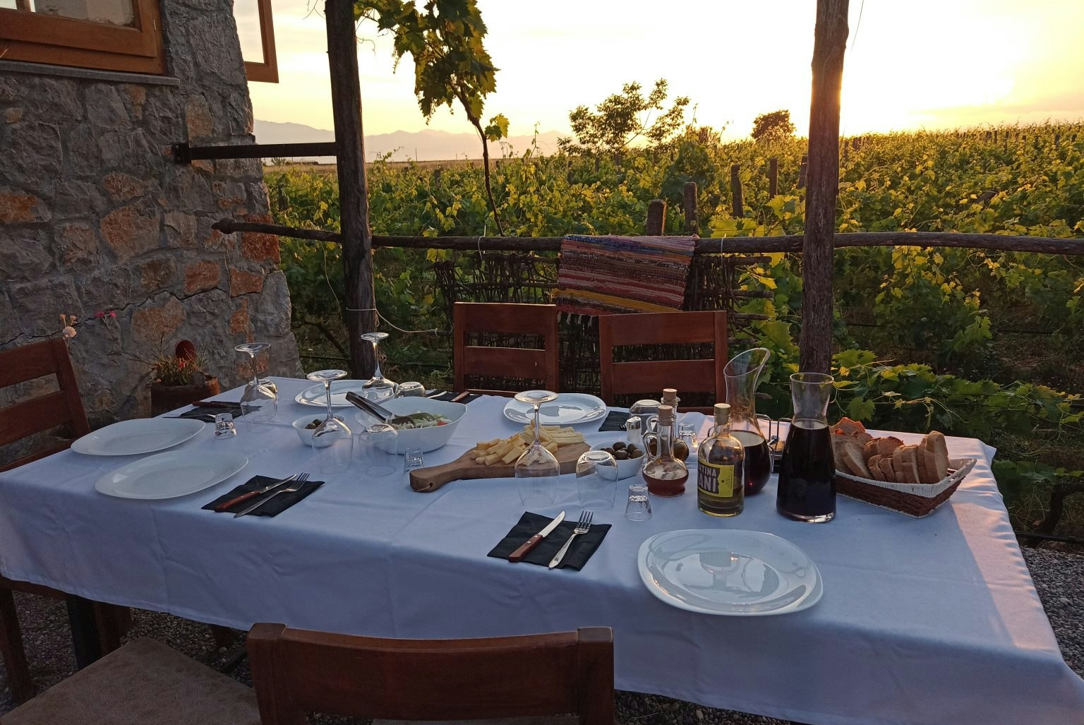 Festlich gedeckter Tisch auf der Camping-Terrasse mit Holzstühlen, Steingartenmauer und Blick in den Garten bei Sonnenuntergang.