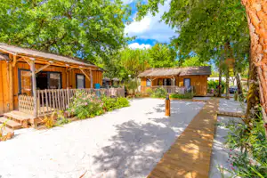 Holz-Bungalows auf einem Campingplatz mit Terrassen, Sitzgelegenheiten und einem Weg zwischen Bäumen und grünem Garten.