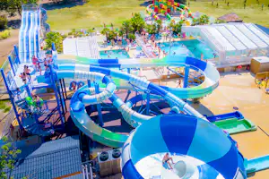 Blaue Wasserrutschen und Kinderbecken auf einem Campingplatz, mit Liegen, Sonnenschirmen und Wegen zwischen Bäumen.