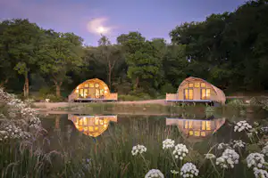 Zwei Glamping-Häuschen mit Holzfenstern am Teich, Spiegelung im Wasser, umgeben von Bäumen und Blumen im Vordergrund.