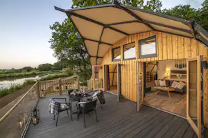 Holzbungalow mit überdachter Terrasse, Gartenmöbeln und Sonnenschirm, Blick auf den Weg und Bäume am Rand des Geländes.