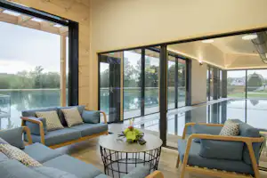 Wohnbereich in einem Mobilheim mit Sofa, Sesseln und Couchtisch auf der Terrasse, große Fensterfront zum See.