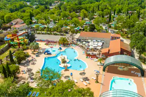 Großer Campingplatz mit mehreren Pools, Liegen und Sonnenschirmen, umgeben von Bäumen und kleinen Gebäuden.