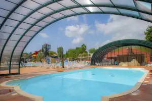 Überdachtes Schwimmbecken auf einem Campingplatz, mit Liegen und Beckenrand, im Hintergrund Wasserattraktionen und Bäume.