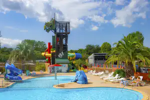 Camping-Pool mit blauen Liegen, Palmen und einem Spielbereich mit Rutsche und Wasserfontäne.
