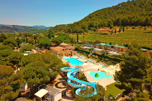 Panoramablick auf einen Campingplatz mit mehreren Pools, Sonnenterrassen und Wohngebäuden inmitten von grünen Hügeln und Bäumen.