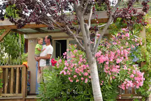 Vor einem Campinghaus stehen zwei Personen am Eingang, umgeben von blühenden Bäumen und Sträuchern im Garten.