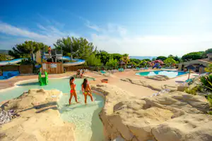 Poolbereich mit Sandboden, mehreren Liegen und Sonnenschirmen, im Hintergrund Bäume und ein Holzdeck.