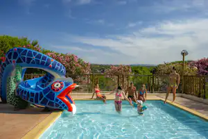 Kinder spielen im Pool vor einer bunten Wasserrutsche, im Hintergrund sind Terrasse, Zäune und grüne Hügel zu sehen.