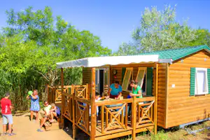 Holzbungalow mit Veranda, Geländer und Sitzbereich; Kinder stehen davor im Grünen mit Bäumen und Weg.