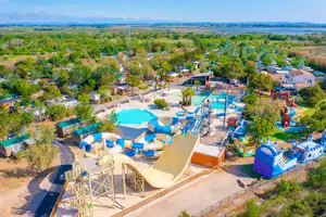 Luftaufnahme eines Campingplatzes mit Pool, Liegen, Sonnenschirmen und Wegen zwischen Stellplätzen im Grünen.