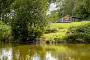Haus am Flussufer auf einer grünen Wiese, umgeben von Bäumen, mit ruhigem Wasser im Vordergrund.