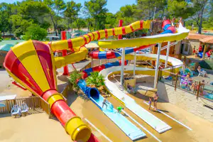 Bunte Wasserrutsche und Pool auf dem Campingplatz, mit Liegen, Sonnenschirmen und Wegen zwischen den Gebäuden.