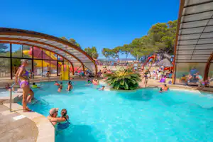 Familien-Pool im Campingbereich mit Sonnenschirmen, Pflanzen und Menschen im Wasser unter einem Glasdach.