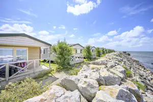 Reihen von Ferienhäusern auf Holzterrassen am Campingplatz, dahinter Sträucher und eine Steinmauer am Meer.