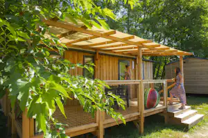 Holz-Terrasse mit Überdachung, Geländer und Stufen am Campingplatz, umrahmt von grünen Bäumen und Sträuchern.