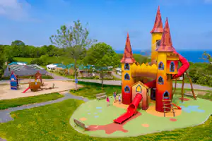 Bunter Spielplatz mit Rutsche und Kletterturm auf dem Campingplatz, im Hintergrund Bäume und das Meer.