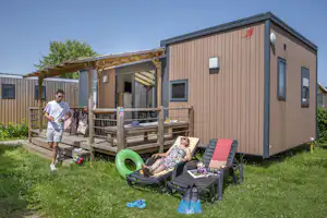 Mobilheim mit Terrasse und Holzgeländer, davor Liegestühle und Rasenfläche; im Hintergrund ein Zaun und weitere Gebäude.