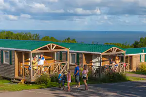 Reihen von Camping-Unterkünften mit Veranda und Holzgeländer, davor ein Weg und Personen, im Hintergrund das Meer.
