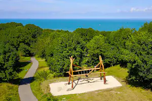 Campingplatz mit kleinem Spielbereich auf einer Wiese, umgeben von Bäumen, mit Blick auf das Meer in der Ferne.