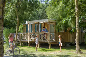 Holzbungalow mit überdachter Terrasse im Grünen, zwei Personen davor, Fahrräder im Hintergrund zwischen Bäumen.
