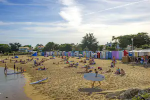 Sandstrand mit vielen Liegestühlen und Sonnenschirmen, im Hintergrund bunte Strandhütten und Bäume, am Rand das Meer.