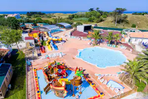 Campinganlage mit beheiztem Pool, Rutschen und Spielbereich, umgeben von Palmen, Bungalows und Blick auf das Meer.