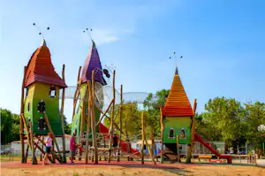 Buntes Spielgerät mit Türmen und Rutschen auf einem Campingplatz, umgeben von Bäumen und blauem Himmel.