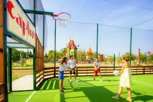 Kinder spielen auf dem grünen Spielbereich mit Basketballkorb und Torwand am Campingplatz, im Hintergrund Holzspielgeräte.