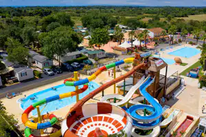 Wasserrutschen und Kinderbecken am Campingplatz, daneben Poolbereich mit Liegen, Bungalows und Bäumen im Hintergrund.