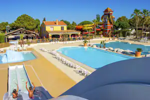 Blick auf den Campingplatz mit zwei Pools, Liegen am Beckenrand und einem gelben Gebäude mit Turm im Hintergrund.