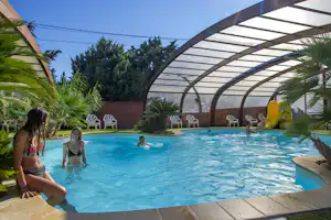 Überdachter Campingpool mit blauem Wasser, Palmen und Liegen; im Hintergrund stehen weitere Sitzgelegenheiten und Bäume.