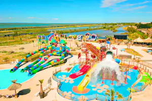Wasserrutschen und Kinderbecken im Campingbereich, mit bunten Spielgeräten, Sonnenschirmen und Blick auf Wasser und Landschaft.