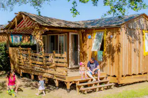 Holzhütte mit Veranda, Sitzbank und Spielzeug; Kinder laufen am Eingang entlang, im Hintergrund Bäume und blauer Himmel.